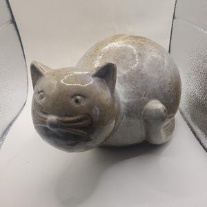 May include: A ceramic cat figurine with a rounded body and a glossy, speckled beige and white glaze. The cat has pointed ears, a round face with defined whiskers, and a smooth, curved posture, showcasing a handcrafted, glazed pottery finish.