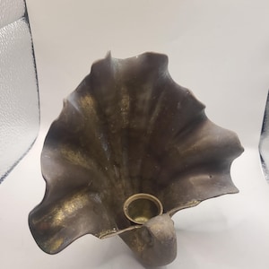 May include: A brass candle holder shaped like a large, open seashell with a dark, weathered patina and a polished gold-toned circular cup at the base for holding a candle. The shell features prominent, radiating ridges and a ruffled, wavy edge.