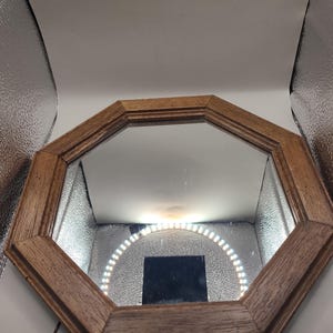 May include: An octagonal wall mirror featuring a medium-toned wooden frame with a visible grain pattern. The reflective glass surface shows a bright circular ring light and a dark square object reflected within the center of the mirror's frame.