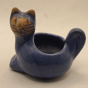 May include: A ceramic cat-shaped planter in a seated position. The planter is primarily blue with a tan face and ears. The open bowl is designed to hold small plants or decorative items. The cat has a whimsical, handcrafted appearance.