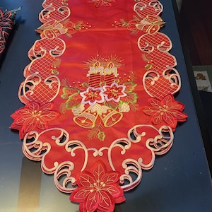 May include: Red Christmas table runner with embroidered poinsettia flowers, bells, and candles. The runner has a scalloped edge and is made of a satin-like fabric.