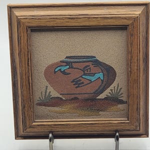 May include: A framed sand painting of a brown pot with blue and white designs, set against a tan background with green plants.