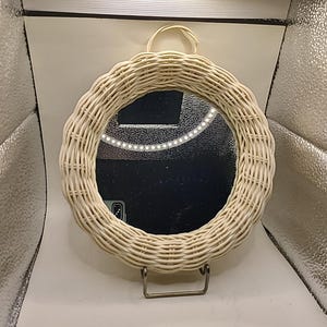 May include: A round, woven wicker mirror with a scalloped edge. The mirror is supported by a metal stand and has a loop for hanging. The mirror's frame is a light beige color.