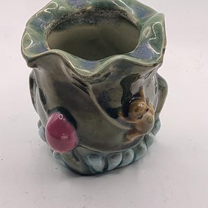 May include: A small green ceramic planter with a frog and a pink mushroom design. The planter has a wavy rim and a textured surface.