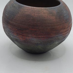 May include: A handcrafted, round ceramic vase with a wide opening. The vase features a textured surface with a gradient of colors, including brown, red, and dark gray. The overall shape is spherical, suitable for home decor.