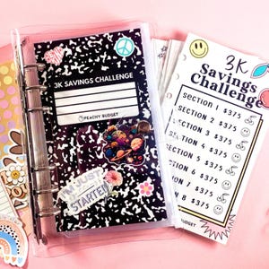 May include: A clear binder with a black and white composition notebook cover that reads "3K Savings Challenge." The notebook is decorated with stickers. A matching savings tracker is visible, with sections for saving $375 each.