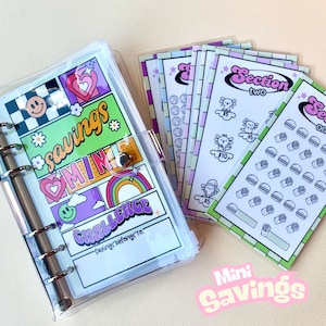May include: A clear plastic binder with a colorful design featuring the words "savings mini challenge". The binder has a section for a name and includes a set of colorful stickers with various designs.