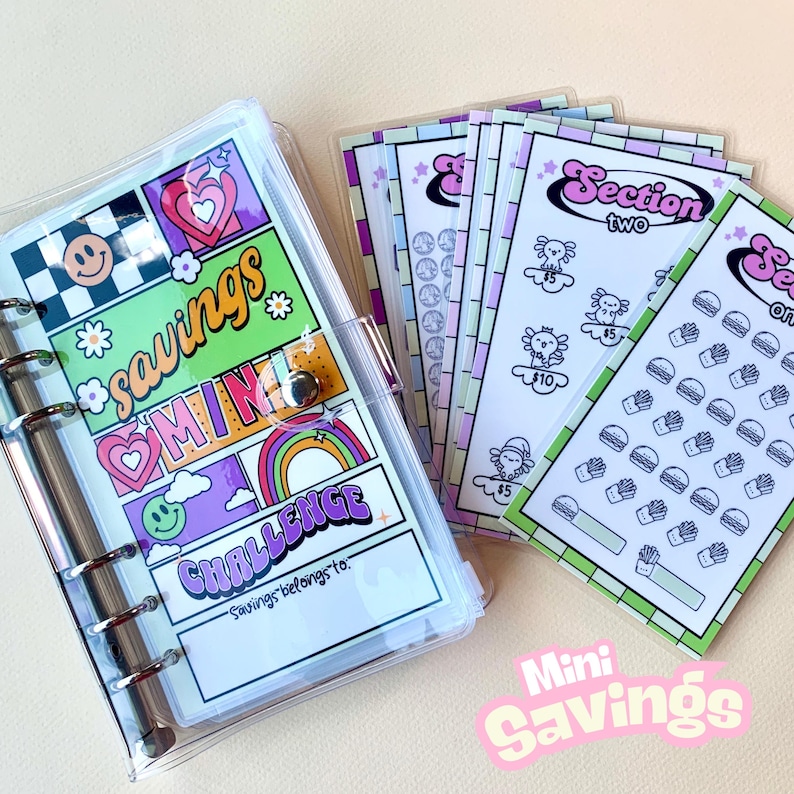 May include: A clear plastic binder with a colorful design featuring the words "savings mini challenge". The binder has a section for a name and includes a set of colorful stickers with various designs.