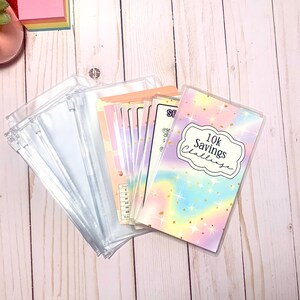 May include: A collection of clear, zippered pouches and colorful budget envelopes. The envelopes feature pastel designs and text, including "10K Savings Challenge." The items are arranged on a light wood surface.