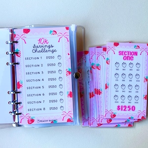 May include: A pink and white planner with a strawberry theme. The planner has a section for a 10k savings challenge with a goal of $1250 per section. The planner also has a section for tracking savings with a goal of $1250.