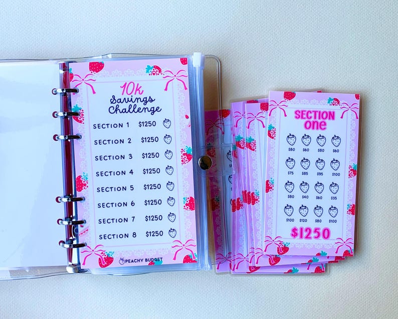 May include: A pink and white planner with a strawberry theme. The planner has a section for a 10k savings challenge with a goal of $1250 per section. The planner also has a section for tracking savings with a goal of $1250.