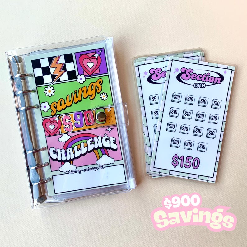 May include: A clear plastic binder with a colorful savings challenge tracker. The tracker features a rainbow, hearts, and the text "$900 Savings Challenge". The binder also includes a set of cards with the text "Section One" and a grid of dollar amounts, totaling $150.