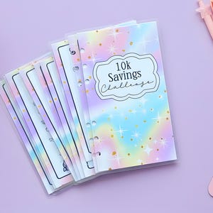 May include: A set of clear plastic budget binders with a pastel rainbow design and gold star accents. The front binder cover has the text "10k Savings Challenge" in a white frame. Pens and a mouse pad are visible.
