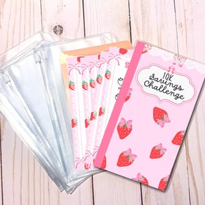 May include: Pink and white savings challenge tracker with a strawberry design and the text "10K Savings Challenge". The tracker is next to a stack of clear plastic zippered pouches.