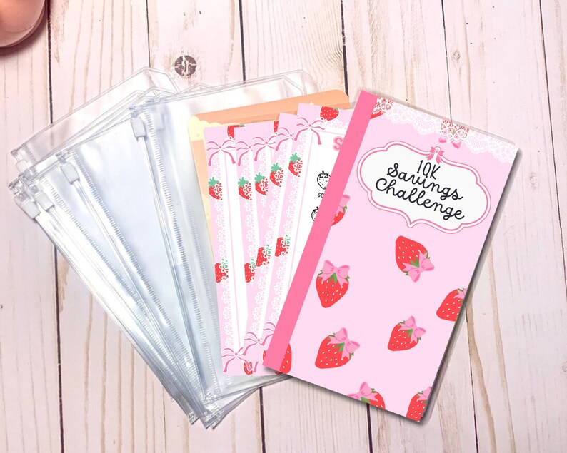 May include: Pink and white savings challenge tracker with a strawberry design and the text "10K Savings Challenge". The tracker is next to a stack of clear plastic zippered pouches.