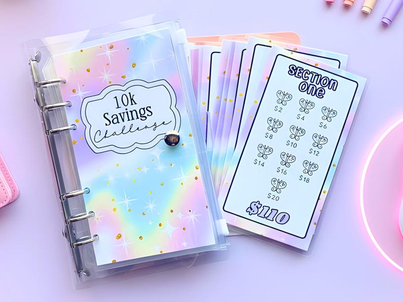 May include: A clear binder with a pastel rainbow cover and the text "10k Savings Challenge." Inside are several savings tracker sheets with butterfly illustrations and amounts from $2 to $20. The total savings amount is $110.