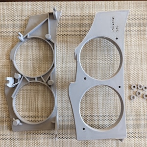 May include: Two light gray automotive headlight housings with circular openings, and a set of small, round bearings. One housing has the text "Porsche 936". The parts are on a textured surface.