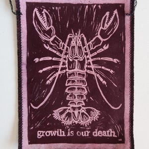 Peut inclure: Écusson en tissu bordeaux et rose avec une illustration de homard blanc. Le patch a une bordure rose et le texte "growth is our death" en bas. Des boucles de fil noir sont aux coins.