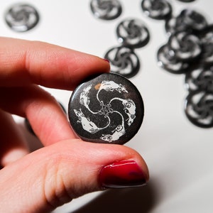 Könnte beinhalten: Ein schwarz-weißer Knopf mit einem Design aus drei stilisierten Fischen, die in einem Kreis schwimmen. Die Fische sind nach innen gerichtet und ihre Schwänze sind eingerollt.