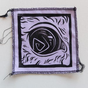 Op de afbeelding: Een vierkante, lavendelkleurige stoffen patch met een zwarte linosnede van een oog. Het oogontwerp heeft een gestileerde pupil en omringende details. De randen zijn afgewerkt met een donkerpaarse gestikte rand en losse draden.