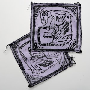 Puede incluir: Dos parches de tela púrpura con bordes cosidos negros. Cada parche presenta una ilustración estilizada en blanco y negro de una criatura con una cola larga y un cuerpo enroscado. La criatura está rodeada de un marco negro.