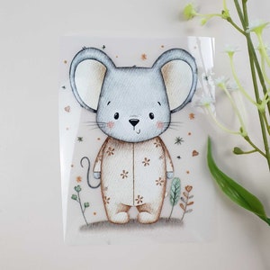 Könnte beinhalten: Eine transparente Transferfolie mit einer Buntstift-Illustration einer grauen Maus mit großen Ohren, die einen beigen Strampler mit kleinen braunen Blumen trägt. Die Maus ist von winzigen orangefarbenen Sternen, Herzen und grünem Blattwerk umgeben.