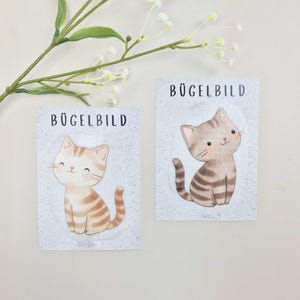 Könnte beinhalten: Zwei rechteckige Karten mit der Aufschrift BÜGELBILD, die jeweils eine Cartoon-Katzenillustration zeigen. Die linke Karte zeigt eine hellorange getigerte Katze, die rechte eine graue getigerte Katze. Beide Katzen haben große Augen und kleine Nasen.