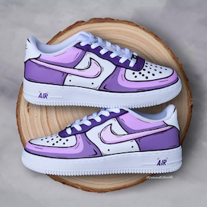 Peut inclure: Paire de baskets Air Force 1 personnalisées. Les chaussures présentent une base blanche avec des accents violets et roses, soulignés de noir. Le mot "AIR" est imprimé sur la semelle intermédiaire. Les baskets sont présentées sur une surface en bois.