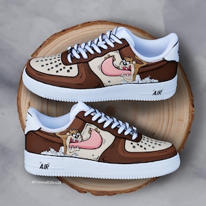 Può includere: Un paio di scarpe da ginnastica dipinte su misura con il personaggio dei cartoni animati Taz, il Diavolo della Tasmania. Le scarpe hanno una combinazione di colori marrone, beige e bianco, con l'immagine del personaggio sul lato. La parola "AIR" è stampata sulla suola bianca.