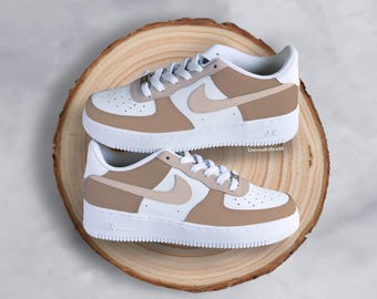 May include: A pair of white and tan sneakers with white laces and a white sole. The sneakers are displayed on a round wooden surface. The Nike swoosh is visible on the side of each shoe. The word "AIR" is printed on the side of the sole.