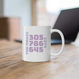 Puede incluir: Taza de cerámica blanca con un diseño rosa y azul. La taza presenta el texto "THE MAGIC CITY" y los números "305, 786, 645".