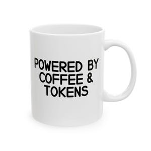 May include: A white ceramic mug featuring the black text "POWERED BY COFFEE & TOKENS" printed in a bold, rounded sans-serif font on the front. The mug has a smooth, glossy finish, a cylindrical body, and a curved handle on the right side.