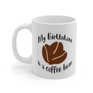 Peut inclure: Une tasse à café en céramique blanche avec du texte noir qui dit "My Birthstone is a coffee bean" et un graphique de trois grains de café.