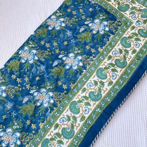 May include: A blue floral patterned table runner. The runner features a repeating pattern of white and blue flowers, green leaves, and yellow accents on a blue background. The border has a white background with green and blue floral designs.