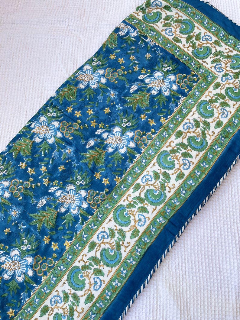 May include: A blue floral patterned table runner. The runner features a repeating pattern of white and blue flowers, green leaves, and yellow accents on a blue background. The border has a white background with green and blue floral designs.