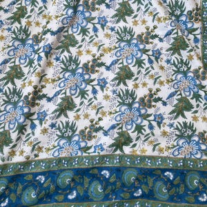 May include: A white fabric with a repeating floral pattern in blue, green, and yellow. The design features large blue flowers with green leaves and smaller yellow flowers. A blue border with a floral design runs along the edge.
