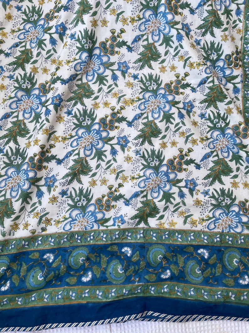 May include: A white fabric with a repeating floral pattern in blue, green, and yellow. The design features large blue flowers with green leaves and smaller yellow flowers. A blue border with a floral design runs along the edge.