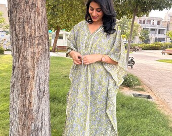 May include: A long, flowing, V-neck kaftan dress with a floral pattern in shades of green, blue, and white. The dress has wide sleeves and a decorative border. The person is standing outside near a tree and green grass.