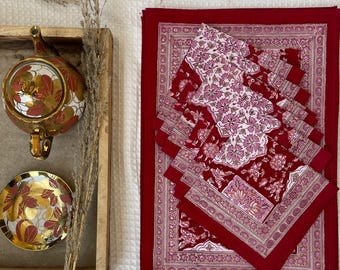 Op de afbeelding: Set van zes rode en witte katoenen placemats met een bloemenpatroon. De placemats zijn gevouwen en gestapeld op een wit oppervlak.