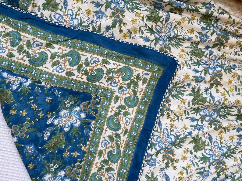 May include: A floral patterned textile featuring a blue border and a white background. The design includes blue, green, and yellow flowers and foliage. The border is a solid blue with a white trim.