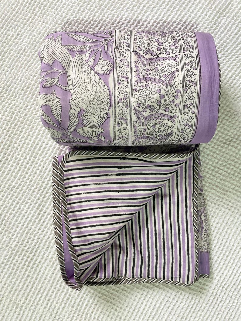 Op de afbeelding: Een gevouwen lavendelkleurige en witte quilt. E&eacute;n kant heeft een vogel- en bloemenpatroon, de andere zwart-witte strepen. Het heeft een grijze rand. De quilt is opgevouwen.