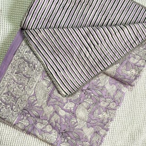 Op de afbeelding: Een lavendel- en witgepatroneerde quilt met een gestreepte achterkant. De quilt heeft een bloemen- en vogelontwerp aan de voorkant en een zwart-wit gestreept patroon aan de achterkant. De randen zijn afgewerkt met een grijze rand.