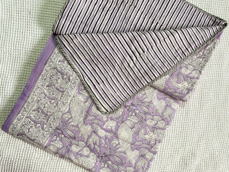 Op de afbeelding: Een lavendel- en witgepatroneerde quilt met een gestreepte achterkant. De quilt heeft een bloemen- en vogelontwerp aan de voorkant en een zwart-wit gestreept patroon aan de achterkant. De randen zijn afgewerkt met een grijze rand.
