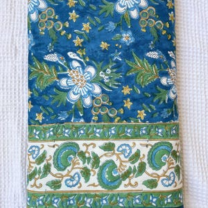 May include: A folded textile with a vibrant floral pattern. The design features large white and blue flowers, green leaves, and small yellow accents on a deep blue background. A white border with green and blue floral motifs is at the bottom.