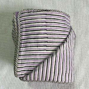 Op de afbeelding: Gevouwen textiel met een gestreept patroon in lavendel, zwart en wit. De randen zijn afgewerkt met een grijs-witte gestreepte rand. De stof is netjes gevouwen en toont het ontwerp.