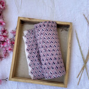 Puede incluir: Dos piezas de tela enrolladas con un estampado floral en una bandeja de madera. La tela es de color malva con un dise&ntilde;o floral p&uacute;rpura oscuro repetido. Tambi&eacute;n se ve una tela blanca con un borde festoneado.