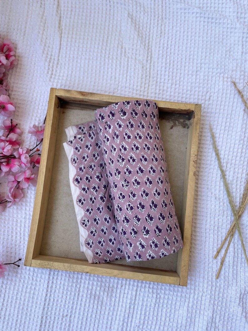 Puede incluir: Dos piezas de tela enrolladas con un estampado floral en una bandeja de madera. La tela es de color malva con un dise&ntilde;o floral p&uacute;rpura oscuro repetido. Tambi&eacute;n se ve una tela blanca con un borde festoneado.