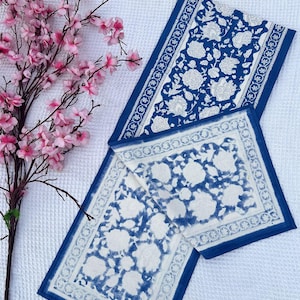 May include: Two blue and white floral patterned table runners and a matching placemat are displayed on a white textured surface. A branch of pink flowers is in the upper left corner. The runners and placemat have a dark blue border.