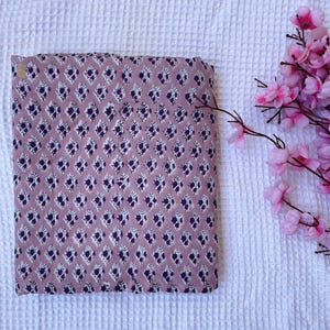 Puede incluir: Tela doblada con un patr&oacute;n repetido de formas florales moradas oscuras y blancas sobre un fondo malva. La tela rectangular parece ser de algod&oacute;n. Flores rosas en la esquina superior derecha.