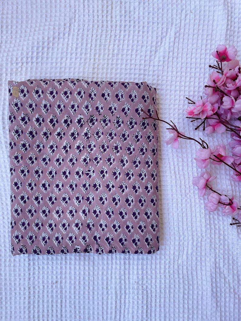 Puede incluir: Tela doblada con un patr&oacute;n repetido de formas florales moradas oscuras y blancas sobre un fondo malva. La tela rectangular parece ser de algod&oacute;n. Flores rosas en la esquina superior derecha.